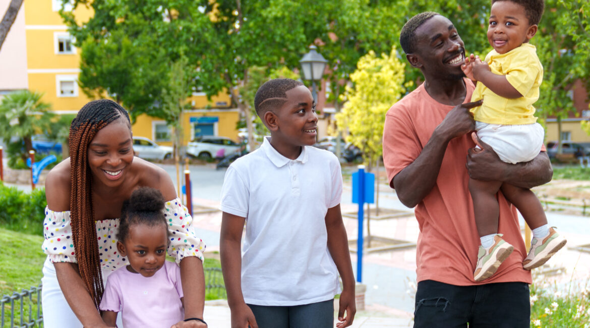 Deux adultes et trois enfants dans un parc : une femme accompagne une petite fille sur une trottinette et un homme porte un jeune enfant.