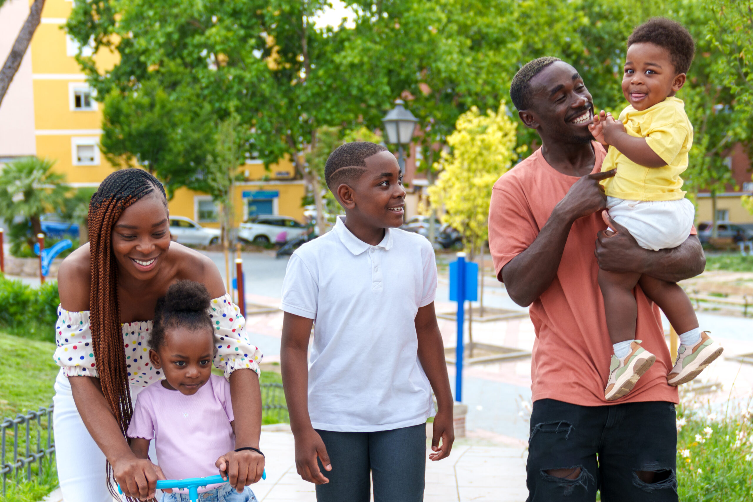 Deux adultes et trois enfants dans un parc : une femme accompagne une petite fille sur une trottinette et un homme porte un jeune enfant.